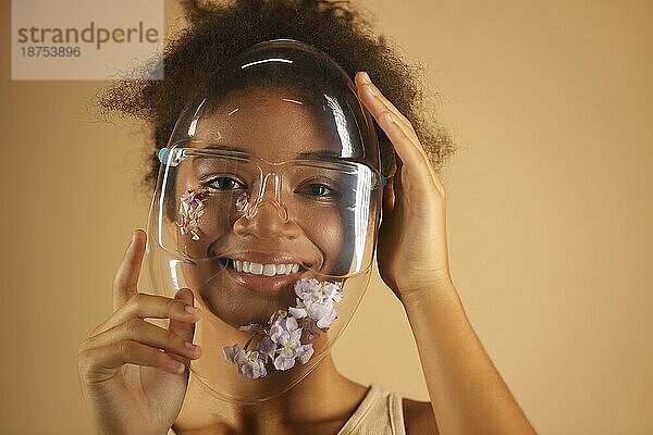Fröhliche afroamerikanische junge Frau  die einen transparenten PVC Schutzschild trägt  der mit Blumen verziert ist und mit einem strahlenden Lächeln in die Kamera blickt  vor einer beigen Wand