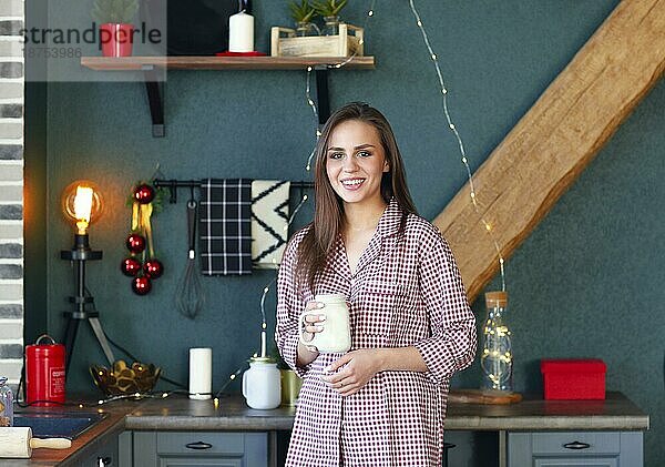 Glückliche junge Frau im karierten Pyjama  die in der weihnachtlich dekorierten Küche ein Glas hält und in die Kamera lächelt