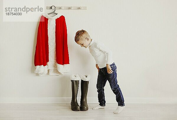 Kleine niedliche Junge Blick auf Santa Claus Kostüm hängen an Holzwand in der Nähe von Schuhen in hellen Raum. Saison celebraion cincept. Winterurlaub