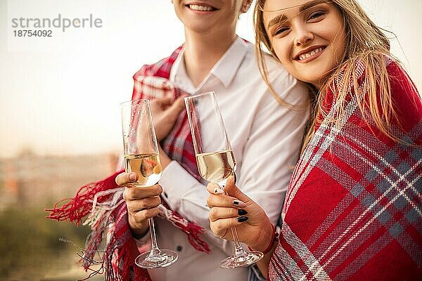 Schönes liebendes Paar  das in eine Decke eingewickelt ist und in die Kamera lächelt  junger Mann und Frau  die Gläser mit Champagner halten  während sie die gemeinsame Zeit bei der Hochzeit von Freunden draußen am Sommerabend genießen