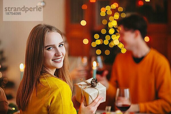 Glücklich lächelnde blonde Frau  die einem Freund ein eingepacktes Geschenk gibt  während sie am Tisch sitzt und gemeinsam Weihnachten feiert