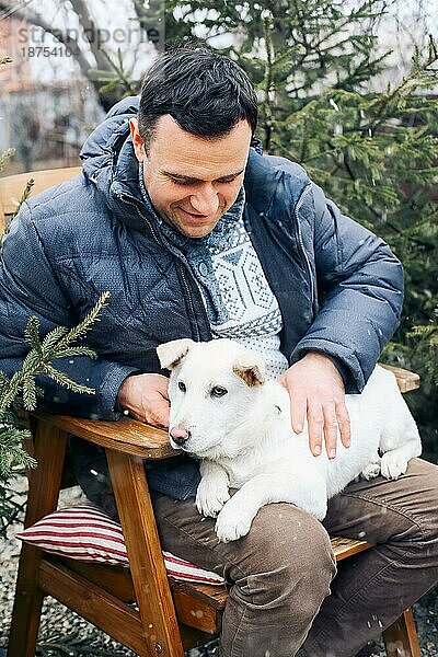 Mann und süßer weißer Hund sitzen auf Stühlen im Park an einem sonnigen Wintertag. Konzept der Freundschaft zwischen Tier und Mensch