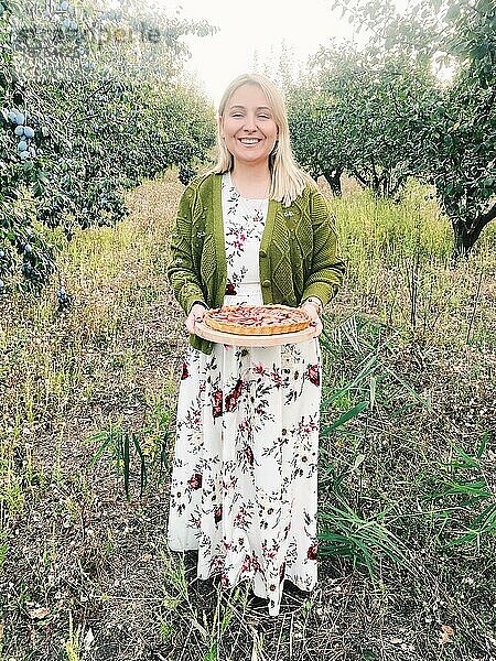 Blonde glückliche Frau in Kleid und Strickjacke trägt Brett mit leckerem Kuchen auf unscharfem Hintergrund der Natur
