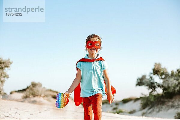 Glückliches Superheldenkind  das am Strand läuft und ein Pop It Spielzeug in der Hand hält