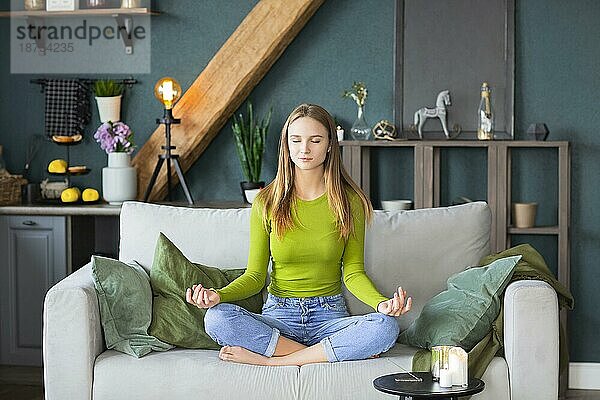 Entspannte friedliche Millennial blondes Mädchen mit geschlossenen Augen sitzen in Lotuspose auf der Couch im Wohnzimmer zu Hause  wobei Pause für Meditation Yoga Praxis. Stressabbau und psychische Gesundheit Konzept