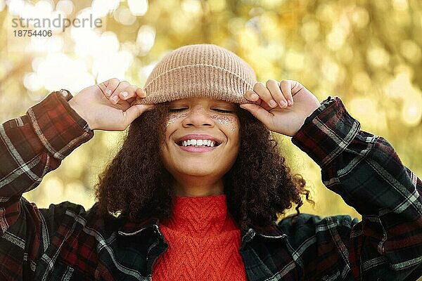 Junge  glückliche  überglückliche afroamerikanische Frau mit lockigem Haar  die ihre beigefarbene Strickmütze zurechtrückt und lacht  während sie im Herbstwald steht  sorglose  gemischtrassige Frau  die die herbstliche Natur im Park genießt
