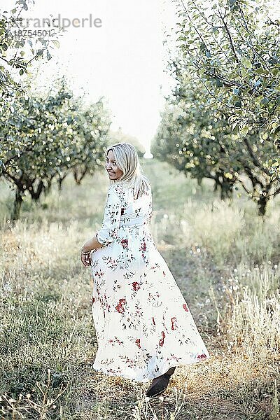 Vertikale Aufnahme einer glücklichen  jungen  sorglosen Frau in einem langen  romantischen Kleid  die in einem grünen Pflaumengarten spazieren geht  über die Schulter in die Kamera schaut und lächelt  während sie an einem Sommertag Zeit in der Natur verbringt. Rückansicht