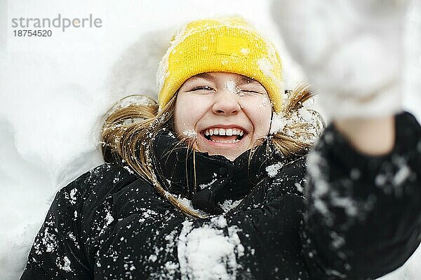 Close up Porträt von lustigen glücklichen Teenagermädchen in gelben Strickmütze spielen mit Schnee im Freien  Kind hält großen Schneeball in der Hand und zeigt es der Kamera. Winter Schnee Spaß für Kinder in der Natur