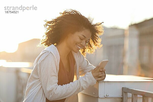 Porträt einer fröhlichen afrikanisch amerikanischen Frau mit Kopfhörern  die ein Smartphone benutzt und glücklich lächelt  während sie auf einem Dach in der Morgensonne steht  fröhliche gemischtrassige Frau mit Mobiltelefon