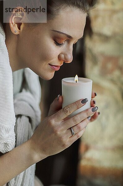 Seitenporträt einer jungen  ruhigen Frau  die eine brennende Kerze in den Händen hält  die Augen schließt und den Duft einatmet. Sie riecht den vertrauten Duft und genießt einen glücklichen Moment des Trostes