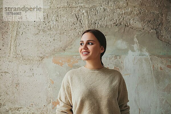 Glückliche junge Frau in beigem Pullover  die lächelnd an einer schäbigen Betonwand steht und den Blick abwendet