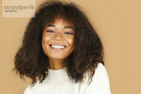 Porträt der wunderschönen fröhlichen afrikanischen amerikanischen jungen Frau mit lockigen Haaren  die in die Kamera lächelt  während sie vor einem beigen Studiohintergrund steht  positive schwarze Frau mit gesunder Haut und zahnigem Lächeln