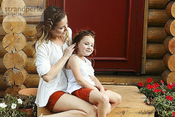 Nettes kleines rothaariges Mädchen und glückliche junge Mutter mit ähnlicher Flechtfrisur umarmen sich  während sie vor einer roten Tür stehen
