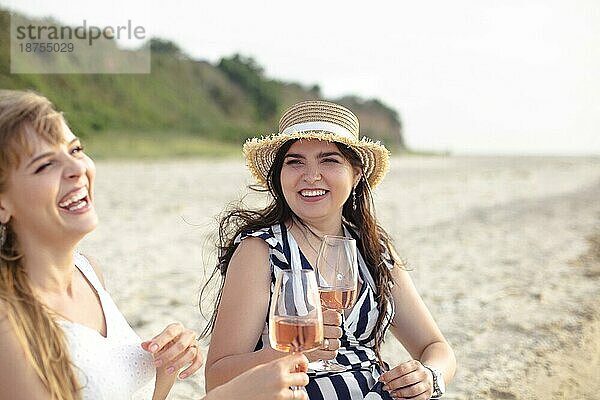 Fröhliche erwachsene Freundinnen in Sommerkleidung lächelnd beim Genießen von Wein am Sandstrand am Meer