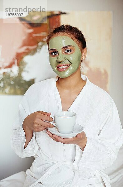 Zufriedene Frau im Bademantel und mit grüner Tonmaske steht mit einer Tasse Tee und genießt das Wochenende im Wellness Center  während sie in die Kamera schaut
