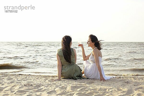 Fröhliche erwachsene Freundinnen in Sommerkleidung lächelnd beim Genießen von Wein am Sandstrand am Meer