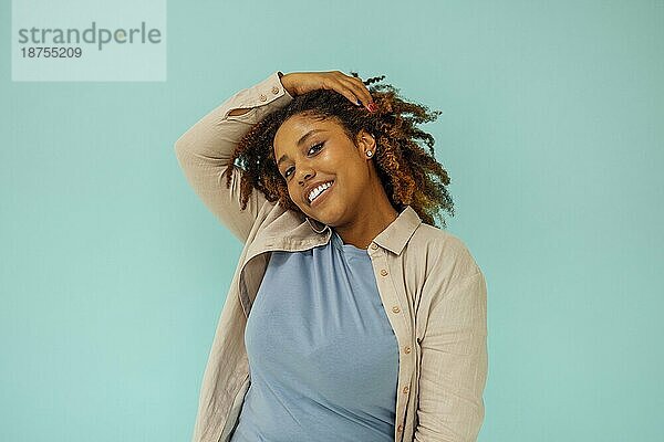 Positives afrikanisches amerikanisches weibliches Modell  das in die Kamera lächelt und sauberes lockiges Haar nach der Hygienebehandlung auf blauem Hintergrund berührt