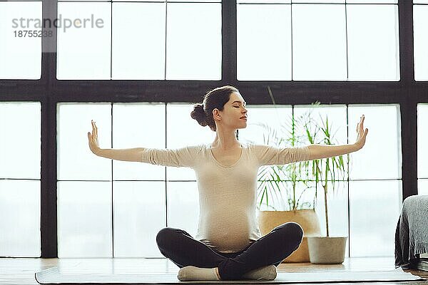 Meditation während der Schwangerschaft. Junge ruhige ruhige schwangere Frau macht Yoga zu Hause  meditieren und üben Achtsamkeit  werdende Mutter sitzt in Lotuspose mit geschlossenen Augen und hob die Arme nach oben
