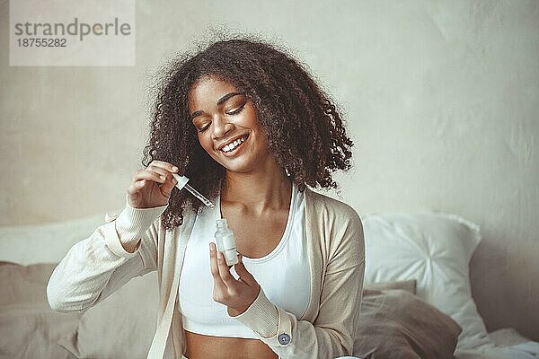 Schönheit und Hautpflege Konzept. Cropped Foto von afrikanischen amerikanischen jungen weiblichen Modell hält Flasche mit feuchtigkeitsspendende Essenz Serum sitzt gegen unscharfen modernen Wohnung Innenhintergrund