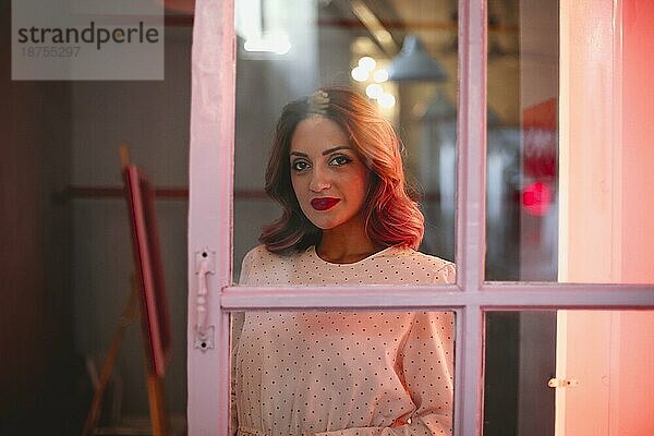 Charmante Frau mit gewelltem Haar steht in der Nähe der Tür in einem von rotem Licht beleuchteten Raum und schaut in die Kamera