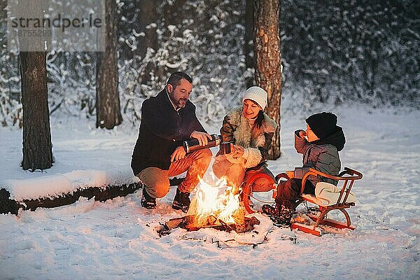 Lächelnde Eltern und Kind braten Marshmallows über dem Lagerfeuer  während sie im Winterwochenende Zeit in der Natur verbringen