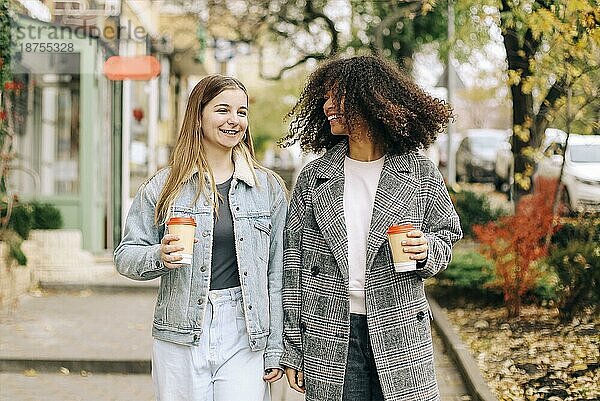 Zwei glückliche sorglose multirassische Freundinnen  die mit einem Kaffee zum Mitnehmen durch die Stadt spazieren  sich unterhalten und Lebensgeschichten austauschen  multiethnische beste Freundinnen  die einen Herbstspaziergang im Freien genießen Freundschaft zwischen Frauen