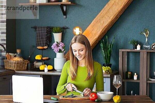 Glückliche Frau lächelt und schneidet Gurke für gesunden Gemüsesalat  während sie das Rezept auf dem Laptop in der Küche zu Hause beobachtet