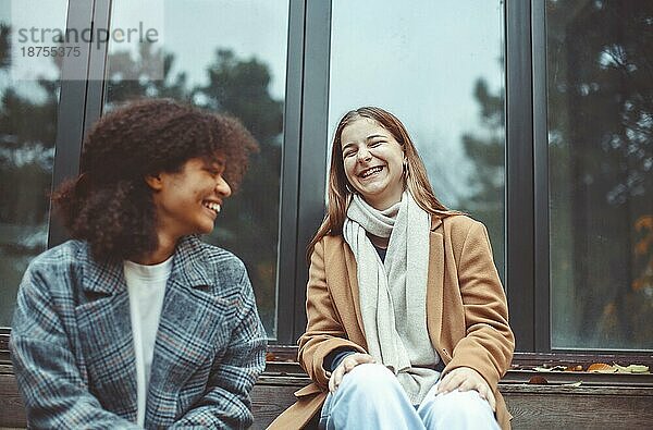 Wo glücklich multirassischen Teenagermädchen mit Kaffee zum Mitnehmen sitzen im Freien auf Herbst Tag  reden und lachen. Lächelnde multiethnische beste weibliche Freunde verbringen Zeit zusammen an der frischen Luft im Herbst Zeit