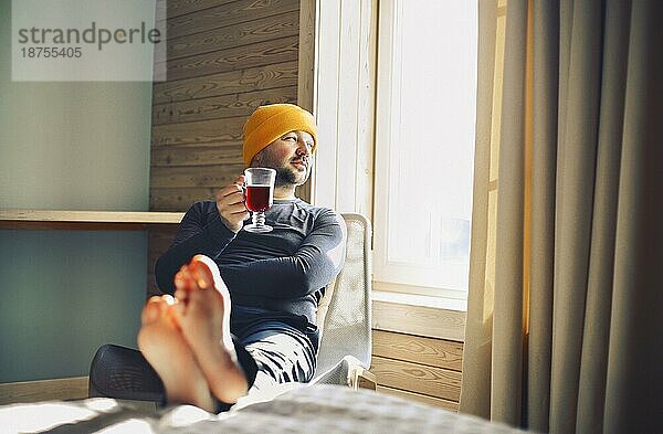 Winter männlichen Reisenden in gelben Hut hält einen Becher Glühwein mit durch das Fenster in hölzernen gemütliches Haus. Männchen trinkt Glühwein im sonnigen Zimmer