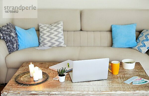 Interieur eines zeitgenössischen Wohnzimmers mit gemütlichem Sofa und Holztisch mit Laptop und Papieren in der Nähe von brennenden Kerzen und einer Tasse Kaffee während der Arbeit von zu Hause aus