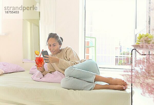 Hübsche junge asiatische Frau in Freizeitkleidung trinkt Fruchtcocktail und surft im Internet auf ihrem Handy  chattet mit Freunden online  während sie auf dem Boden im gebadeten Sonnenlicht Zimmer zu Hause sitzt