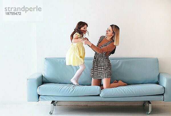 Glückliches verspieltes kleines Mädchen Tochter spielt mit positiven Mutter  Springen auf dem Sofa  verbringen Zeit zusammen während der Ferien zu Hause  Mama und Kind Spaß haben im Wohnzimmer