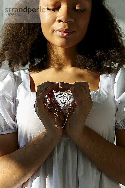 Junge friedliche dankbare afroamerikanische Frau mit geschlossenen Augen in weißem Kleid macht Herz Zeichen  führte Lichter in den Händen der ruhigen schwarzen Frau zeigt Symbol der Liebe. Nächstenliebe Konzept