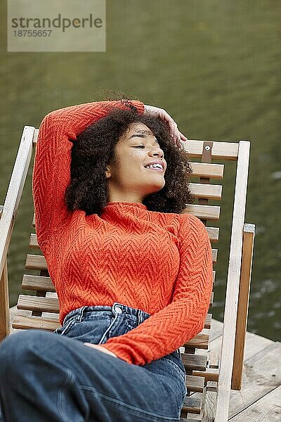 Junge glückliche entspannte afrikanische amerikanische Frau mit lockigen Haaren in orange warmen Pullover entspannende Ruhe auf Liegestuhl in der Nähe von Teich oder Fluss im Herbst Wochenende Tag. Menschen und Erholung in der Natur