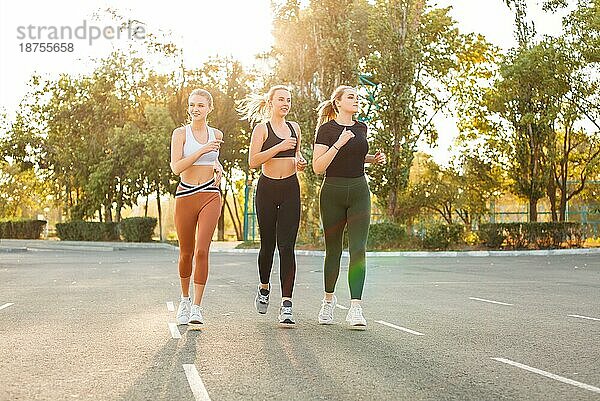 Ganzkörper junge Sportlerinnen lächelnd und laufen auf Asphalt Parkplatz auf sonnigen Tag im Park