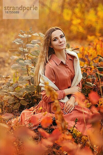 Schöne junge blonde Frau trägt Boho Stil langes Kleid beim Sitzen mit nachdenklichen Blick in hohem Gras im Herbst Wald in roten und gelben Farben dekoriert  Blick weg mit verträumten geheimnisvollen Lächeln