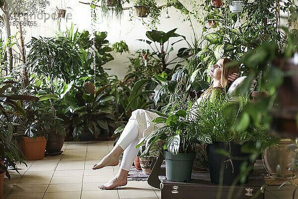 Ruhige Frau mit Händen hinter dem Kopf auf einem Stuhl in einem Raum voller Blumentöpfe mit üppigen Pflanzen liegend