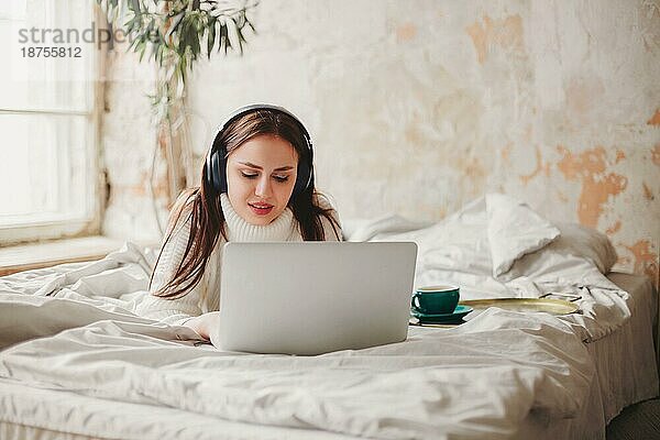 Junge Frau hört Musik und surft in den sozialen Medien auf einem Netbook  während sie sich morgens auf dem Bett bei einer Tasse Kaffee ausruht