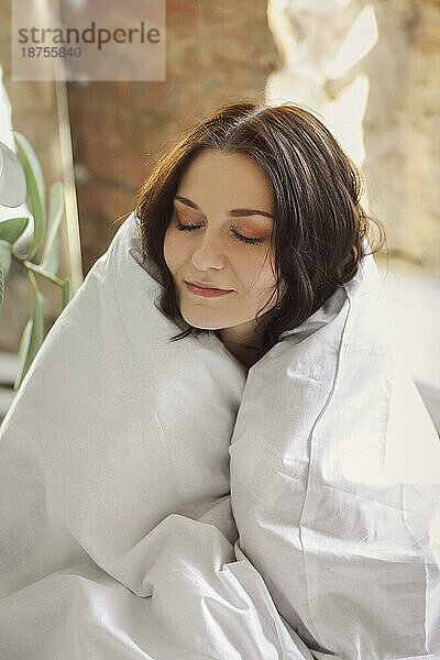 Porträt einer jungen  schönen  glücklichen Frau  die in eine Bettdecke eingewickelt sitzt  die Augen schließt und vor Freude und Komfort lächelt  während sie am frühen sonnigen Morgen in einer gemütlichen  heimeligen Atmosphäre aufwacht