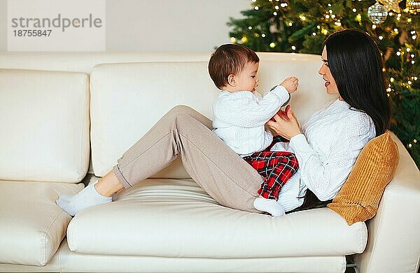 Junge Frau lächelt und spielt mit süßem Jungen auf unscharfem Hintergrund von geschmücktem Nadelbaum am Weihnachtstag zu Hause