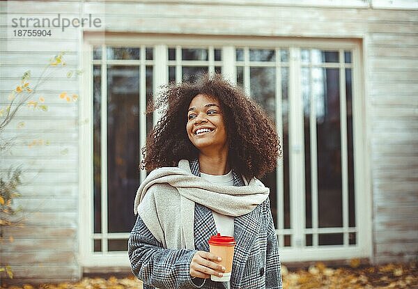 Herbststimmung. Junge glückliche afroamerikanische Frau mit lockigem Haar im Herbst Park hält Kaffee zum Mitnehmen  schaut zur Seite und lächelt  entspannt in der Natur  genießt ihre Lieblingsjahreszeit. Selektiver Fokus