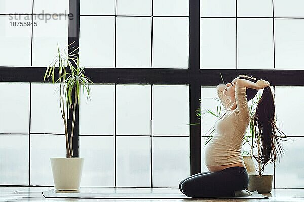 Junge ruhige ruhige schwangere Frau bindet Haare in Pferdeschwanz vor Yoga oder Stretching Praxis zu Hause  werdende Mutter sitzt auf Fitnessmatte gegen große Panoramafenster. Sport und Schwangerschaft