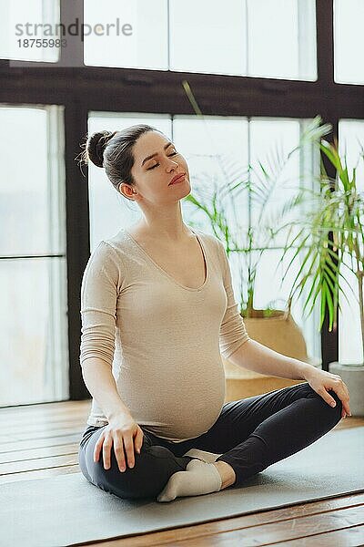 Meditation während der Schwangerschaft. Junge ruhige ruhige schwangere Frau macht Yoga zu Hause  meditieren und üben Achtsamkeit  werdende Mutter sitzt im Lotussitz mit geschlossenen Augen
