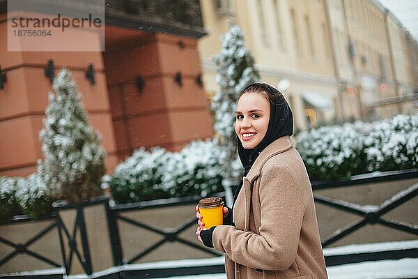 Seitenansicht einer fröhlichen jungen Frau in Oberbekleidung  die lächelt und in die Kamera schaut  während sie einen Kaffee zum Mitnehmen auf einem verschwommenen Hintergrund einer verschneiten Straße im Winter genießt