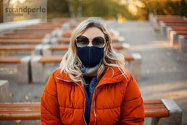 Blonde Frau in Oberbekleidung und Maske geht auf einem gepflasterten Weg an einem sonnigen Tag im Herbstpark während einer Pandemie