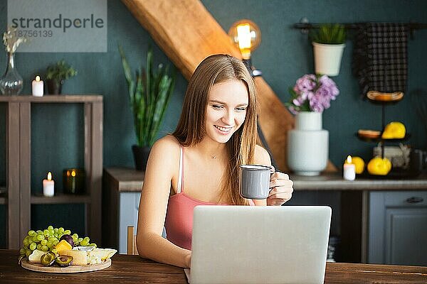 Glückliche junge Frau mit einer Tasse Heißgetränk  die lächelt und am Morgen zu Hause im Laptop surft