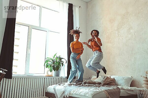 Zwei glückliche  fröhliche  multirassische Freundinnen springen auf dem Bett und haben Spaß zusammen zu Hause  während sie ihre Freizeit verbringen  positive weibliche Freunde verschiedener Rassen  die lachen und die weibliche Freundschaft genießen