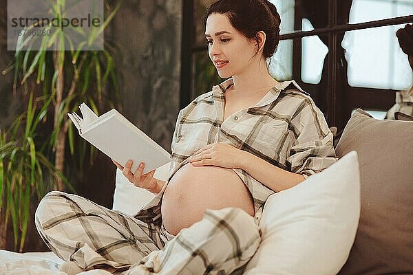 Junge  attraktive  glückliche schwangere Frau in Hauskleidung  die ein Buch liest und ihren Bauch berührt  während sie sich zu Hause auf der Couch ausruht. Die werdende Mutter trägt einen karierten Pyjama und genießt ihre Freizeit während der Schwangerschaft