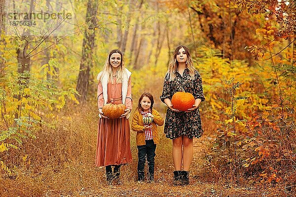 Schuss von Freunden stehen außerhalb mit gruseligen Gesicht Kürbis in den Händen auf dem Weg voll von gefallenen Blättern im Herbst Park  zwei erwachsene Frauen und kleines Mädchen feiern Halloween Urlaub