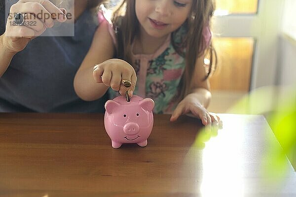 Kleines Mädchen  das eine Münze in ein Sparschwein steckt  während es mit seiner Mutter zu Hause am Tisch sitzt  eine Frau  die ihrem Kind beibringt  wie man vernünftig mit Geld umgeht  indem sie die Grundlagen der Haushaltsführung für Kinder erklärt. Finanzielle Bildung